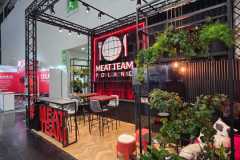 Nowoczesne stoisko targowe "MEAT TEAM POLAND" z czerwono-czarnym wystrojem, zestawem jadalnym, stołkami barowymi, zielonymi roślinami i oznakowaniem marki z logo talerza, widelca i noża. Górne światła oświetlają stoisko, tworząc przyjazną atmosferę. projekt i nadzór Tatjana Kobuszewska trksystem.com