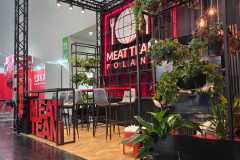 Nowoczesne stoisko wystawiennicze MEAT TEAM POLSKA ma czarne metalowe ramy, bujne zielone rośliny, wiszące światła, drewnianą podłogę i czerwone oznakowanie. Wewnątrz stoiska znajdują się wysokie krzesła i stoły, z widocznym wystrojem MEAT TEAM i elementami wzornictwa przemysłowego. projekt i nadzór Tatjana Kobuszewska trksystem.com