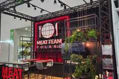 Nowoczesne stoisko targowe z oznakowaniem MEAT TEAM POLSKA z logo widelca i noża, czerwonymi akcentami LED, wysokimi stołkami przy drewnianej ladzie, roślinami doniczkowymi i dekoracją z sylwetką czarnej świni. Reflektory nad głową oświetlają stylową, industrialną aranżację. projekt i nadzór Tatjana Kobuszewska trksystem.com
