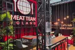 Nowoczesne stoisko wystawiennicze z czarno-czerwonym wystrojem, z napisem "MEAT TEAM POLAND" i logo globu na ścianie. Wysokie krzesła i stoły, wiszące lampy, zielone rośliny i czerwony napis MEATTEAM wypełniają stylowo zaprojektowaną przestrzeń. projekt i nadzór Tatjana Kobuszewska trksystem.com