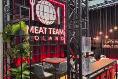 Nowoczesne stoisko wystawiennicze z napisem MEAT TEAM POLAND na czerwono podświetlonej ścianie z logo kuli ziemskiej, widelca i noża. Wysokie stoły z szarymi krzesłami, rośliny doniczkowe i wiszące lampy tworzą stylową, zachęcającą atmosferę. projekt i nadzór Tatjana Kobuszewska trksystem.com