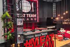 Nowoczesne stoisko targowe z odważnymi czerwonymi literami "MEATTEAM" z przodu, ladą z krzesłami, roślinami i czarną ścianą z dużym podświetlanym logo Meat Team Thailand. Stoisko jest otoczone czarną metalową konstrukcją, ma drewnianą podłogę i przytulne siedzenia. projekt i nadzór Tatjana Kobuszewska trksystem.com