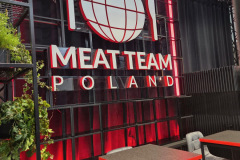 Restauracja ze stolikami i krzesłami przed dużym napisem MEAT TEAM POLAND z symbolem kuli ziemskiej, widelca i noża, całość podświetlona czerwonym światłem. Zielone rośliny zdobią lewą stronę obrazu. projekt i nadzór Tatjana Kobuszewska trksystem.com