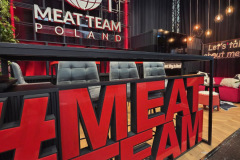 Nowoczesne stoisko z dużymi czerwonymi literami "MEAT TEAM", trzema szarymi krzesłami i świecącym napisem "MEAT TEAM POLAND" z kulą ziemską, widelcem i nożem. Ciepłe wiszące światła i strefa wypoczynkowa tworzą przyjazną atmosferę. projekt i nadzór Tatjana Kobuszewska trksystem.com
