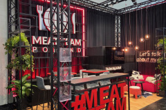 Nowoczesne stoisko wystawiennicze z czarną metalową ramą i czerwonymi neonami zawiera podświetlany napis "MEAT TEAM POLAND", ladę z dużym czerwonym napisem MEATTEAM, zieleń, lampy wiszące, czarne krzesła i czerwoną kanapę, tworząc żywą, przyjazną atmosferę. projekt i nadzór Tatjana Kobuszewska trksystem.com