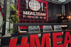 Na stoisku targowym widnieje duży napis MEAT TEAM POLSKA z logo kuli ziemskiej, widelca i noża na czarno-czerwonym tle. Czarne krzesła są ustawione przy ladzie, a odważny czerwony napis MEAT TEAM zdobi pierwszy plan. Zielone rośliny dodają odrobinę koloru. projekt i nadzór Tatjana Kobuszewska trksystem.com