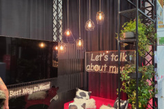 Przytulna budka z czerwoną kanapą, poduszkami ze skóry bydlęcej, małymi czarnymi stolikami i wiszącymi lampami wiszącymi. Neony z napisami "Porozmawiajmy o mięsie" i "Najlepszy sposób na mięso". W rogu znajdują się zielone rośliny, a w tle czarne zasłony i półki. projekt i nadzór Tatjana Kobuszewska trksystem.com