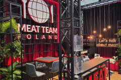 Nowoczesne stoisko wystawiennicze z dużym napisem "MEAT TEAM POLAND", czerwono-czarnym wystrojem, wysokimi stołami z szarymi stołkami barowymi, zielonymi roślinami, wiszącymi lampami i ścianą z napisem "Porozmawiajmy o mięsie" świecącymi literami. Stoisko ma metalowe ramy w stylu industrialnym. projekt i nadzór Tatjana Kobuszewska trksystem.com