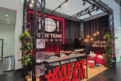 Nowoczesne stoisko wystawowe z czarną ramą i czerwonymi akcentami prezentuje branding "MEAT TEAM". Na stoisku znajdują się rośliny, oświetlenie punktowe, leżaki i lada. Z przodu znajduje się duży czerwony znak "MEAT TEAM", a podświetlany napis głosi "Porozmawiajmy o mięsie". projekt i nadzór Tatjana Kobuszewska trksystem.com