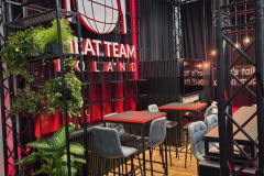 Nowoczesna kawiarnia z wysokimi stołami i szarymi stołkami barowymi, czarnymi metalowymi konstrukcjami, zielonymi roślinami doniczkowymi i biało-czerwonym napisem "CAT TEAM POLAND" na ścianie. Ciepłe wiszące światła i drewniana podłoga dodają przytulnej atmosfery. projekt i nadzór Tatjana Kobuszewska trksystem.com