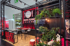 Nowoczesne stoisko targowe z czarną ramą, czerwonymi akcentami i napisem "MEAT TEAM POLAND". Na stoisku znajdują się stołki barowe, stoły, wiszące rośliny, odsłonięte żarówki i napis MEATTEAM. Zieleń zdobi przestrzeń, kontrastując z czystym, industrialnym designem. projekt i nadzór Tatjana Kobuszewska trksystem.com