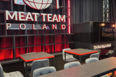 Nowoczesne wnętrze restauracji z napisem "MEAT TEAM POLAND" pogrubionymi czerwonymi i białymi literami na ścianie. Nad tekstem znajdują się duże ikony widelca, noża i kuli ziemskiej. Są szare krzesła, czarne stoły i ciemny, industrialny wystrój z czerwonym oświetleniem akcentującym. projekt i nadzór Tatjana Kobuszewska trksystem.com