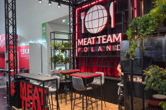 Nowoczesne stoisko targowe dla Meat Team Poland ma czarną metalową ramę, jaskrawoczerwony neon, wysokie stoły z szarymi stołkami, zielone rośliny i podłogę z wzorem drewna. Z przodu znajduje się duży czerwony napis 3D z hashtagiem MEAT TEAM. projekt i nadzór Tatjana Kobuszewska trksystem.com