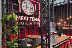 Nowoczesne stoisko wystawiennicze z napisem MEAT TEAM POLAND na czerwono-czarnej ścianie, z ikoną kuli ziemskiej, widelca i noża. Stołki barowe, wysoki stół, zielona ściana z roślinami, geometryczne światła i drewniana podłoga. projekt i nadzór Tatjana Kobuszewska trksystem.com
