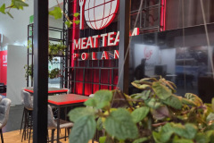 Kryte stoisko z czerwono-białym napisem MEAT TEAM POLAND z logo kuli ziemskiej i widelca. Nowoczesne stołki barowe i stoły stoją na drewnianej podłodze, a zielone rośliny na pierwszym planie dodają scenie odrobinę natury. projekt i nadzór Tatjana Kobuszewska trksystem.com