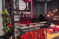 Nowoczesne stoisko wystawowe z dużym czerwonym napisem "MEAT TEAM" z przodu, elegancką czarną metalową ramą, drewnianą podłogą, siedzeniami barowymi, czerwonym oświetleniem neonowym, logo o tematyce mięsnej na tylnej ścianie, roślinami i telewizorem. Przestrzeń jest stylowa i zachęcająca. projekt i nadzór Tatjana Kobuszewska trksystem.com