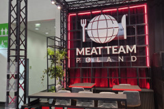 Stoisko targowe z metalową ramą i neonami wyświetla napis "MEAT TEAM POLAND" pod logo talerza, widelca i noża. Z przodu znajduje się duży czerwony hashtag "MEATTEAM" wraz z ladą, stołkami i roślinami doniczkowymi. projekt i nadzór Tatjana Kobuszewska trksystem.com