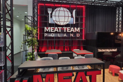 Nowoczesne stoisko wystawiennicze z odważnymi czerwonymi literami MEAT TEAM z przodu, czarną ladą ze stołkami, roślinami po lewej stronie i dużym podświetlanym napisem "MEAT TEAM POLAND" z kulą ziemską, widelcem i nożem na tylnej ścianie. projekt i nadzór Tatjana Kobuszewska trksystem.com