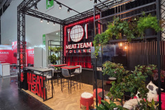 Nowoczesne stoisko wystawiennicze Meat Team Poland charakteryzuje się czerwono-czarnym wystrojem, roślinami, stołami, krzesłami i podświetlanymi znakami z naczyniami. Widoczna jest niewielka część wypoczynkowa ze stołkami i poduszkami, a także wiszące rośliny i drewniana podłoga. projekt i nadzór Tatjana Kobuszewska trksystem.com