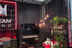 Nowoczesny salon z czerwoną sofą, małymi czarnymi stolikami i szarymi krzesłami na drewnianej podłodze. Wiszące lampy, zielone rośliny i czarne tło z podświetlanym oznakowaniem i czerwonymi akcentami tworzą przytulną, stylową atmosferę. Czarna metalowa rama wyznacza przestrzeń. projekt i nadzór Tatjana Kobuszewska trksystem.com