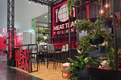 Nowoczesne stoisko wystawowe z napisem MEAT TEAM POLSKA ma czarne metalowe ramy, drewnianą podłogę, bujne zielone rośliny, wiszące światła i szare krzesła wokół stołów. Odważne czerwone litery oznaczają MEATTEAM przy wejściu. W tle widoczne są znaki i inne stoiska. projekt i nadzór Tatjana Kobuszewska trksystem.com