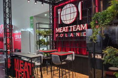 Nowoczesne stoisko targowe MEAT TEAM POLSKA obejmuje czarne metalowe ramy, czerwono-czarną kolorystykę, logo ścienne z widelcem, nożem i kulą ziemską, wysokie stoły z szarymi stołkami, rośliny doniczkowe i odważne oznakowanie MEATTEAM na drewnianej podłodze. projekt i nadzór Tatjana Kobuszewska trksystem.com