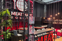 Nowoczesne stoisko targowe zawiera odważne czerwone akcenty, czarne ramy, duży napis "MEAT TEAM" z logo kuli ziemskiej i widelca, neonowy napis "Porozmawiajmy o mięsie", miejsca do siedzenia, rośliny i bar, tworząc stylową i przyjazną atmosferę. projekt i nadzór Tatjana Kobuszewska trksystem.com