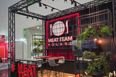 Nowoczesne stoisko wystawiennicze z czarną metalową ramą i czerwonymi akcentami prezentuje napis MEAT TEAM POLAND z logo kuli ziemskiej, widelca i noża. Dekoracyjne rośliny, stołki barowe i drewniana podłoga tworzą przyjazną atmosferę. MEAT TEAM wyróżnia się pogrubionymi czerwonymi literami z przodu. projekt i nadzór Tatjana Kobuszewska trksystem.com