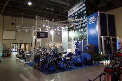 Stoisko promocyjne prezentuje różne niebieskie maszyny i urządzenia do czyszczenia przemysłowego w jasnym świetle w dużej hali wystawowej z podłogami wyłożonymi kafelkami i wysokimi sufitami. Na znakach widnieje napis Nilfisk ALTO. projekt i nadzór Tatjana Kobuszewska trksystem.com