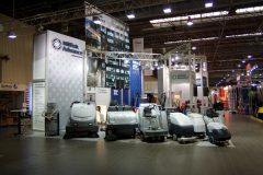 Stoisko targowe prezentujące różne przemysłowe maszyny i urządzenia czyszczące na podłodze wyłożonej kafelkami, z banerami produktowymi i oświetleniem nad głową. projekt i nadzór Tatjana Kobuszewska trksystem.com