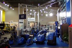 Stoisko targowe prezentuje kilka niebieskich maszyn i urządzeń do czyszczenia przemysłowego w jasnym świetle, ze stoiskami informacyjnymi i uczestnikami w tle. projekt i nadzór Tatjana Kobuszewska trksystem.com