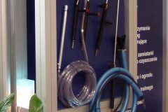 Tablica reklamowa z różnymi urządzeniami czyszczącymi, wężami i narzędziami zamontowana na ścianie, z rośliną doniczkową i podświetlanymi kostkami na podłodze poniżej. projekt i nadzór Tatjana Kobuszewska trksystem.com