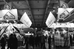 Czarno-białe zdjęcie ruchliwych targów handlowych przedstawia ludzi spacerujących między różnymi stoiskami firmowymi, w tym podwójnym stoiskiem prezentującym materiały dachowe i okna dachowe pod dużymi znakami „Koto”. Jasno oświetlona hala wystawowa tętni życiem. projekt i nadzór Tatjana Kobuszewska trksystem.com