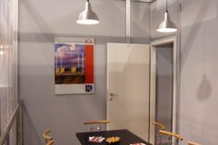 Mała sala konferencyjna z czarnym stołem, sześcioma czerwonymi krzesłami z poduszkami, dwoma wiszącymi lampami, otwartymi białymi drzwiami i oprawionym obrazem na szarej ścianie. Broszury i popielniczki znajdują się na stole w tym podwójnym stoisku. projekt i nadzór Tatjana Kobuszewska trksystem.com