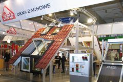 Podwójne stoisko dostarczające okno dachowe oraz świetliki z modelami skośnych dachów, czerwonymi dachówkami, plakatami informacyjnymi i rozmowami odwiedzających. Na miejscu zobacz szyld „OKNA DACHOWE”. projekt i nadzór Tatjana Kobuszewska trksystem.com