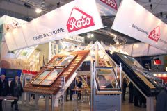 Stoisko targowe, na którym eksponowane są okna dachowe i świetliki marki „Roto” na skośnych pozornych dachach. Nad głowami wiszą banery, a wokół stoiska krąży kilka osób. projekt i nadzór Tatjana Kobuszewska trksystem.com