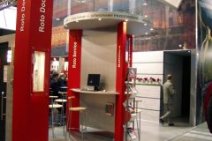 Nowoczesne stoisko podwójne z czerwonymi filarami oznaczonymi „Roto DoorTec” i „Roto Service”, z ladą, stołkami, komputerem i ekspozycją ścienną — idealne do prezentacji produktów takich jak okna dachowe — w ruchliwym centrum kongresowym. Ludzie chodzą w pobliżu i wokół stoiska. projekt i nadzór Tatjana Kobuszewska trksystem.com