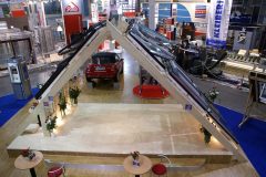 Na stoisku targowym znajduje się duża drewniana konstrukcja w kształcie litery A z wieloma oknami dachowymi Roto Okna Dachowe na targach Budma. Wewnątrz zaparkowany jest czerwony samochód, otoczony kwiatami i miejscami do siedzenia. W tle widoczne są znaki i stoiska. projekt i nadzór Tatjana Kobuszewska trksystem.com