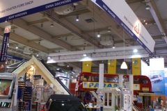 Stoisko targowe z zaparkowanym przed nim czerwonym samochodem, opatrzonym logotypami. Duże wiszące szyldy podkreślają Roto Okna Dachowe na targach Budma, z ekspozycjami okien, drzwi, wnętrza domu i czerwonym piętrowym autobusem w tle. projekt i nadzór Tatjana Kobuszewska trksystem.com