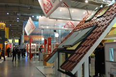 Targi, na których kilka osób spaceruje wśród eksponatów, w tym dużego dachu z czerwonymi dachówkami i oknami dachowymi Roto, pod banerami z logo „Roto” wewnątrz przestronnej, dobrze oświetlonej hali na targach Budma. projekt i nadzór Tatjana Kobuszewska trksystem.com