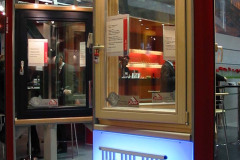 Ekspozycja targowa Roto budma obejmuje dwie pionowe ramy okienne — jedną czarną, jedną z jasnego drewna — z ekspozytorami okiennymi i brandingiem Roto. Przednia rama znajduje się nad świecącym, podświetlonym na niebiesko panelem, a w tle widać ludzi i stoły. projekt i nadzór Tatjana Kobuszewska trksystem.com