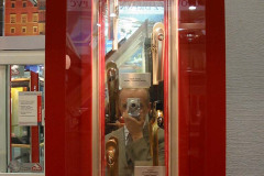 Osoba robiąca zdjęcie odbija się w szklanej gablocie zawierającej wiele metalowych klamek do drzwi, obramowanej czerwoną ramką i jasno oświetlonej — jest to część podwójnego stoiska z górnym oknem dachowym wpuszczającym naturalne światło. projekt i nadzór Tatjana Kobuszewska trksystem.com