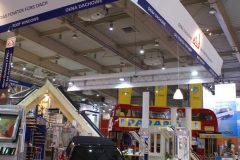 Kryte stoisko wystawowe na targach Budma obejmuje czerwonego Mini Coopera z logo firmy, duże wiszące szyldy, ekspozytory okien i piętrowy autobus w tle – wszystko pod wysokim, industrialnym sufitem. projekt i nadzór Tatjana Kobuszewska trksystem.com