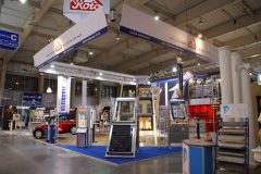 Stoisko targowe Budma eksponuje firmę Roto okna dachowe, różnorodne okna, drzwi i materiały budowlane na niebieskim dywanie, nad głowami wiszą banery, a na oknach ekspozytora w dobrze oświetlonej hali wystawienniczej starannie ułożono próbki produktów. projekt i nadzór Tatjana Kobuszewska trksystem.com