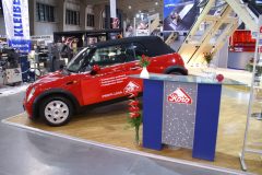 Czerwony kabriolet Mini Cooper, przypominający klasycznego Mini Morrisa, jest wystawiony w pomieszczeniu obok lady ze szklanym blatem i logo Roto. Stoiska wystawowe i banery w tle podkreślają Roto okna dachowe na targach Budma. projekt i nadzór Tatjana Kobuszewska trksystem.com