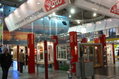 Stoisko targowe z czerwono-białym szyldem Roto i różnymi ekspozycjami okien i drzwi na targach Budma. Ludzie chodzą wokół wystawy, a nad głowami wiszą banery z polskim tekstem „Otwieramy Przestrzenie”. projekt i nadzór Tatjana Kobuszewska trksystem.com