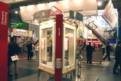 Stoisko targowe prezentuje okna dachowe Roto Frank w jasnym świetle. Kilka osób spaceruje po wystawie ekspozytory okuć okiennych, a nad głowami w krytym centrum kongresowym targi budma wiszą tablice informacyjne. projekt i nadzór Tatjana Kobuszewska trksystem.com