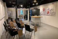 Nowoczesna klasa z czarnymi krzesłami i przymocowanymi drewnianymi ławkami ustawionymi w rzędach. Idealna jako sala dydaktyczna montażu okien Roto Frank, posiada centralne podium, duży ekran, tablicę, mapę na ścianie i przeszklone biuro z tyłu. projekt i nadzór Tatjana Kobuszewska trksystem.com