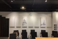 Nowoczesna sala konferencyjna z czarnymi krzesłami ustawionymi w rzędach, drewnianym stołem z przodu, czterema szkicami architektonicznymi na ścianie i okrągłym zegarem nad szkicami tworzy idealną przestrzeń Roto frank sala dydaktyczna montażu okien pod jasnymi światłami sufitowymi. projekt i nadzór Tatjana Kobuszewska trksystem.com