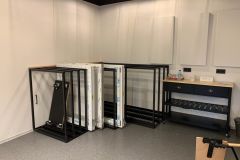 Kilka dużych czarnych metalowych ram i zapakowanych paneli, używanych do Roto frank sala dydaktyczna montażu okien, jest ułożonych na białej ścianie. Po prawej stronie czarny stolik konsolowy zawiera szuflady, wodę butelkowaną i pudełko na herbatę. W pokoju znajduje się szara podłoga. projekt i nadzór Tatjana Kobuszewska trksystem.com